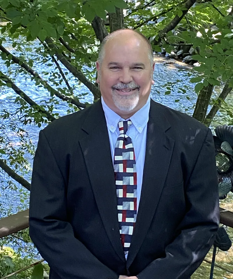 Jim Goodyear in a dark suit and tie, smiling, beside the Chattahoochee River in Helen
